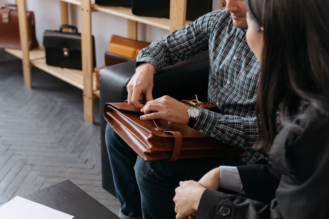 gallery-1 A man demonstrates a brown leather briefcase to a woman in a stylish showroom.