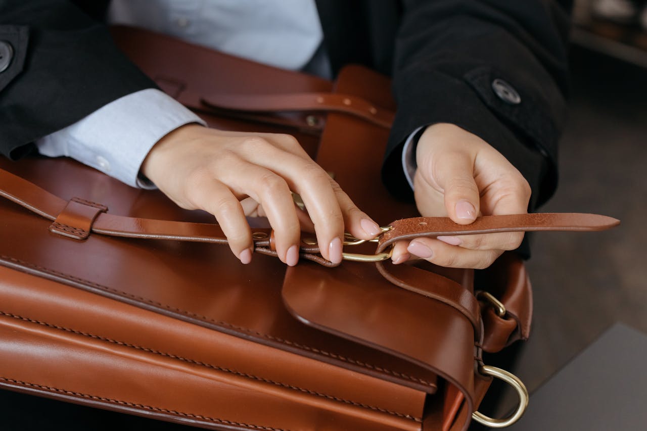 gallery-5 Close-up of hands fastening a brown leather briefcase strap indoors.